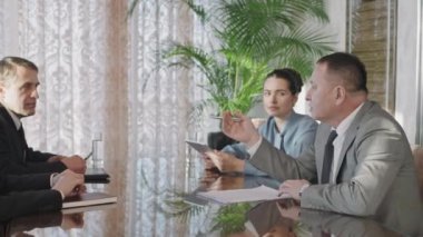 Medium PAN shot of four male and female investors sitting at negotiation table discussing business strategy. Middle-aged Asian man gesturing while speaking to his business partners