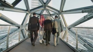 Modern yüksek sınıf ofis binasının cam köprüsü boyunca yürümekte olan çok ırklı erkek ve kadın iş ortaklarından oluşan orta büyüklükte bir yavaşlama.