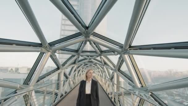 オフィスビルの長い空のガラス橋に沿って歩く黒のスーツで自信を持って中年の実業家のフロントビューのドローンスロモ — ストック動画