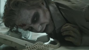 Beyin ölümü gerçekleşmiş SFX makyajlı ve sahte yaraları olan zombi ofis çalışanının masada yatıp telefona cevap vermeye çalışırken yakın plan çekimi.