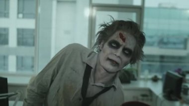 SFX makyajlı, yırtık gömlekli ve kravatlı zombi adamın boş bir ofiste kameraya doğru yürüdüğünü gösteren el kamerası görüntüsü.