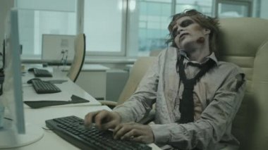 Zombi ofis çalışanının masa başında oturup elleriyle klavyeyi kırdığı orta boy bir fotoğraf.