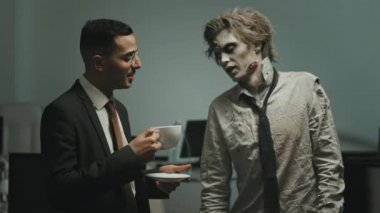 Gözlüklü ve takım elbiseli bir iş adamının kahve içip ofiste yırtık gömlekli ve kravatlı zombi adamla konuştuğu orta boy bir fotoğraf.