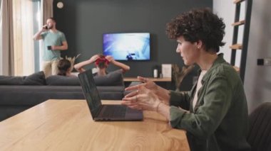 PAN orta ölçekli, kendinden emin bir kadının ev bilgisayarından iş videosu çekilirken kocası pencerenin önünde telefonla konuşurken ve arka planda iki çocuk video oyunu oynarken.