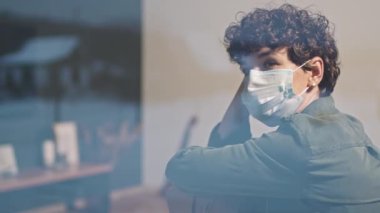 Düşünceli, koruyucu maske takmış beyaz bir kadın, pencerenin kenarında oturmuş, hayatı düşünüyor.