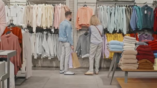衣料品店で一緒に美しい若い喜びのカップルのショッピングのフルショット 女取りトレンディーなスウェットシャツからラックショーへ彼の意見を求める — ストック動画
