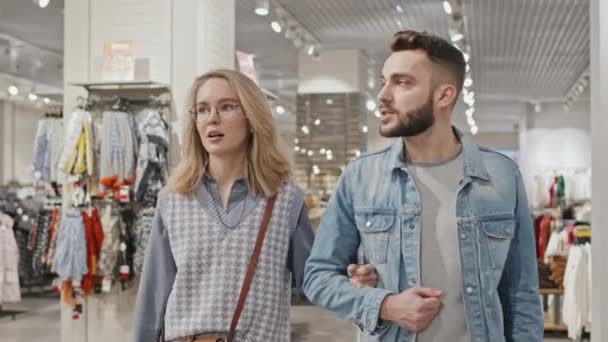 洋服屋さんでカメラショッピングに向かって一緒に歩いている間に話しているスタイリッシュな若い白人カップルの中程度のショット — ストック動画