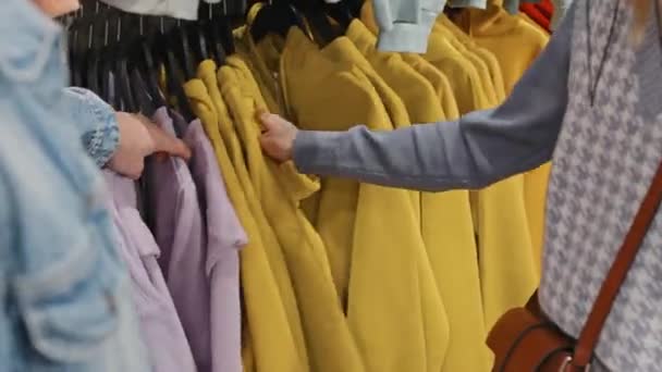 右のサイズを探している衣料品店でレールにかかって鮮やかな色の並べ替えのパーカーの認識できないカップルのクローズアップ — ストック動画
