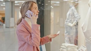 Genç ve çekici bir kadının büyük bir alışveriş merkezinde dikilip şık giyim mağazasının vitrinine bakarken orta boy fotoğrafı.