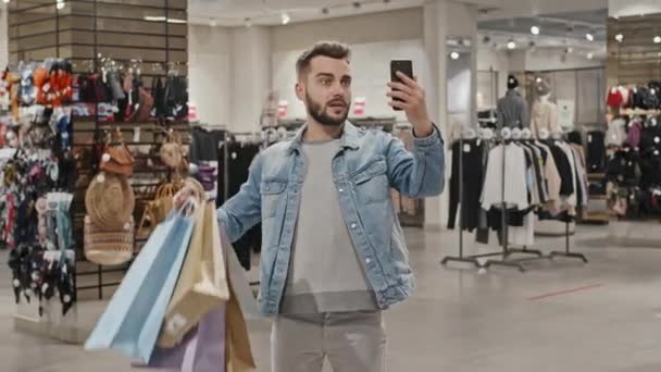 カメラに新しい服で買い物袋の多くを示す衣料品店でビデオ通話を持つ流行のデニムジャケットでハンサムな男の中出し — ストック動画