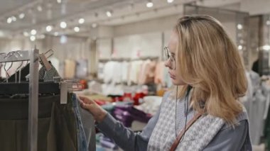 Gözlüklü, çekici, sarışın bir kadının belden yukarı pozu. Kıyafet mağazasında zincirli, pantolon seçen.