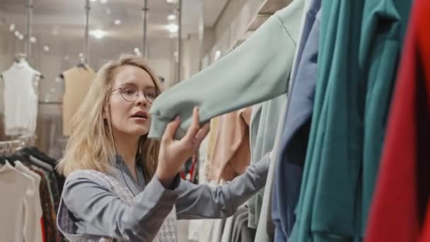 ウェストアップショットの美しい若い女性で流行の眼鏡ショッピングのために服で現代店のソートトレーナーにぶら下がっレール — ストック動画
