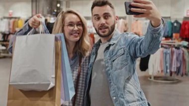 Orta ölçekli mutlu genç çiftin elbise mağazasında görüntülü sohbet ederken fotoğraf çekimi. Bir sürü alışveriş torbasını kamera önünde sohbet ederken ve gülümserken gösteriyor.