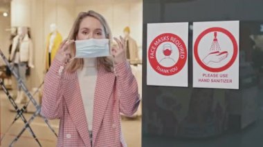 Giysi mağazasının girişinde basılı covid önleyici levhalarla dururken kameraya bakan şık bir kadının orta boy portresi.