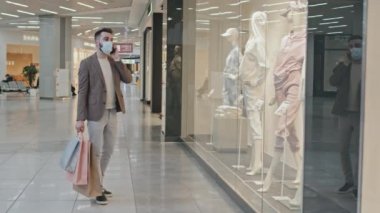 Yakışıklı, sakallı, şık giyinmiş, elinde bir sürü alışveriş çantası olan ve büyük bir alışveriş merkezinin vitrininin önünde telefonla konuşan bir adam.