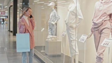 Büyük bir alışveriş merkezinin vitrininde beklerken telefonda gülümseyen, şık giyinmiş güzel bir kadının orta boy fotoğrafı.