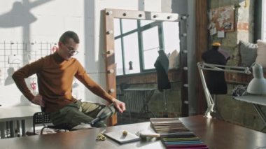 Güneşli bir günde havalı stüdyoda protez bacaklı, sandalyede oturan ve kameraya poz veren şık erkek moda tasarımcısının PAN yavaşlığı.