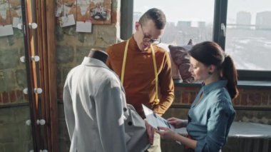 Gözlüklü bir erkek moda tasarımcısının stüdyodaki mankene bakıp kadın terziye kumaş örnekleri gösterirken orta boy fotoğrafı.