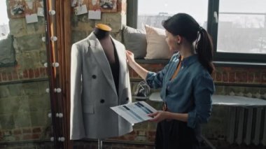 Kadın moda tasarımcısının kumaş örnekleri tutmasının ve mankene asılı duran cekete dokunmasının yavaşlığını izleyip kameraya doğru dönüyor ve gülümsüyor.