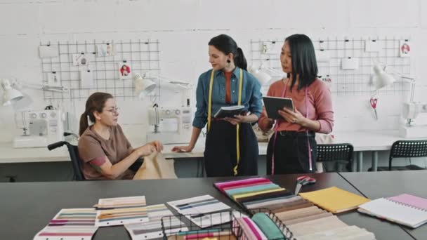 跟踪照片上的快乐女性时装设计师 带着笔记本和平板电脑与轮椅上的女裁缝交谈 给她们展示工作室里的服装 — 图库视频影像