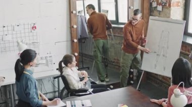 Protez bacaklı erkek moda tasarımcısının üzerine kıyafet çizimi yapılmış beyaz tahtanın önünde dikilirken ve terzilerle toplantı yaparken çekilen fotoğrafı.