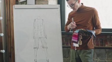 Gözlüklü erkek moda tasarımcısının yavaş yavaş öne eğip kumaş örnekleri tutmasını ve ekibiyle görüşürken giysi fikrini sunmasını istiyorum.