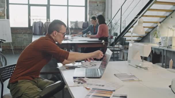 穿着假腿的男性时装设计师坐在办公桌前 一边看着笔记本电脑 一边勾画出新系列的构思 一边慢镜头跟踪镜头 — 图库视频影像