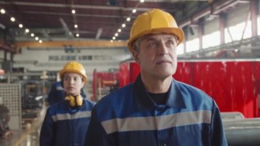 Tulumlu kadın ve erkek işçilerden oluşan belden aşağısı yavaş ve kasklı bir şekilde fabrikadan geçerek etrafa bakınıyorlar. Ciddi bir yetişkin erkek müfettişi devam ediyor.