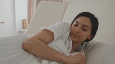 Latin kökenli, beyaz çarşafların üzerine uzanmış, güzel bir uykudan sonra uyanıp esneyen genç ve çekici bir kadının yakın çekimi.