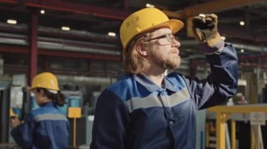 Yavaş sakallı fabrika işçisi üniformasını giymiş güneşe bakıyor ve iş yerinde duran sarı kaskı ayarlıyor.