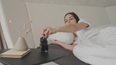 Latin kökenli neşeli genç bir kadın sabah uyanıp çalar saati kapatıyor ve rahat beyaz yatakta uzanırken gülümsüyor.