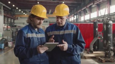 İki fabrika işçisinin kasketli ve üniformalı orta yavaş çalışması fabrika tesisinde dijital tablete bakarak çalışmalarını tartışıyor sonra da kameraya başlarını çeviriyor.