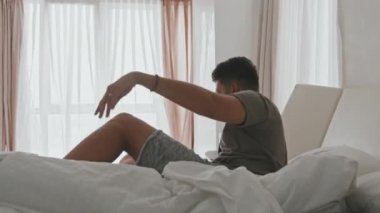 Latin kökenli genç bir adamın sabahları uyanıp panoramik pencereye bakarak rahat bir yatakta uzanırken orta açıdan çekilmiş bir fotoğrafı.