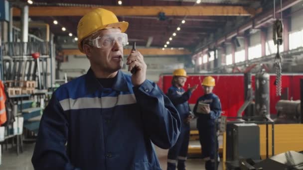 跟踪戴安全眼镜和硬礼帽的成年男性主管在通过工厂设施时的慢动作 并向员工发出对讲机指令 — 图库视频影像