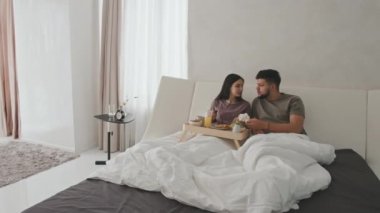 Genç evli bir çiftin panoramik pencereli modern minimalist yatak odasında romantik bir kahvaltı yaparken yatakta oturup konuştuğu geniş bir hava görüntüsü.