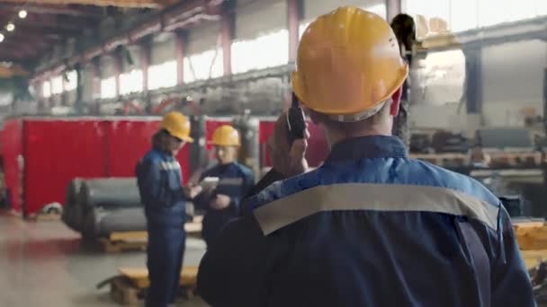 戴着安全眼镜和硬礼帽的男性主管穿过工厂设施时的后视镜慢镜头 并向员工发出对讲机指令 — 图库视频影像