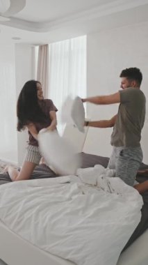 Pijamalı genç Latin bir çiftin yatakta yastık savaşı yaparken çekilmiş dikey bir fotoğrafı.