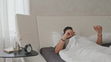 Yakışıklı Latin adamın evde rahat bir yatakta uyanıp çalar saate baktığı ve tembelce esnediği orta boy bir fotoğraf.
