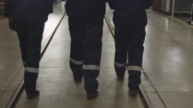 Tilt-up rear-view shot with slowmo of three male and female workers in coverall uniforms and hard hats walking through plant facility during shift