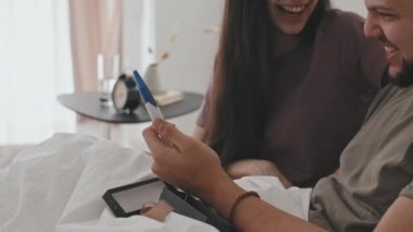 Mutlu, evli, yatakta oturan ve hamilelik testi sonucu pozitif çıkan bir çift. Gülümseyen genç kadın, şok olmuş kocasının tepkisini izliyor.