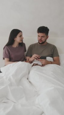 Romantik genç çiftin sabahları rahat bir yatakta otururken hamilelik testine bakarken orta boy görüntüsü. Kocasının hediye kutusuna hamilelik testi koyan bir kadın.