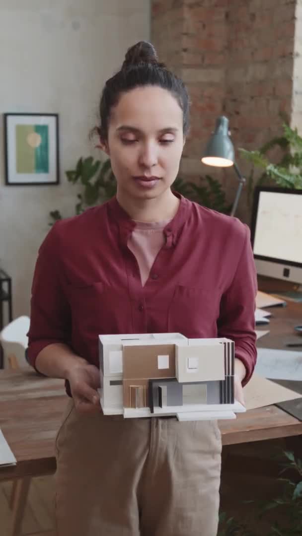 現代のスタイリッシュな建築事務所に立っている3Dペーパーハウスレイアウトを保持カメラを見てプロの女性建築家の垂直メディア肖像画 — ストック動画
