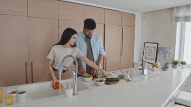 Modern minimalist tarzda mutfakta birlikte yemek pişirirken konuşan gündelik elbiseli genç bir Latin çiftin orta boy fotoğrafı. Kadın taze sebze yıkıyor ve erkek onları kesiyor.