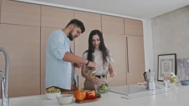 Minimalist parlak mutfakta dikilmiş birlikte sebze salatası yapan gündelik kıyafetli genç bir Latin çiftin orta boy fotoğrafı. Adam zeytin yağı eklerken, kadın malzemeleri kaseye karıştırıyor.