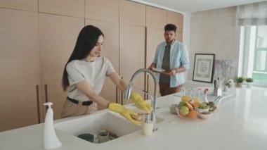 Genç ve mutlu bir Latin erkeğin, güzel karısına bulaşık yıkaması için yardım ederken minimalist tarzda modern tasarım mutfağında onları yıkarken çekilmiş orta boy bir fotoğrafı.