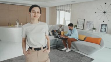 Latin kökenli çekici bir kadının orta boy portresi modern oturma odasında elleri ceplerinde kameraya gülümserken erkek arkadaşı ya da kocası arka planda dizüstü bilgisayarla çalışırken.