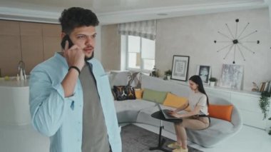 Latin kökenli yakışıklı, vahşi bir adamın arka plandaki karısı modern rahat oturma odasında dizüstü bilgisayarla otururken telefonda konuştuğu orta boy bir fotoğraf.