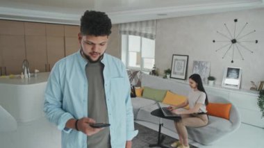 Genç Latin bir adamın akıllı telefon kullanıp kameraya gülümserken karısı modern tasarım oturma odasında dizüstü bilgisayarda çalışırken orta ölçekli görüntüsü.