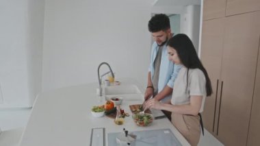 Latin kökenli yakışıklı genç bir adamın modern minimalist mutfakta yan yana dururken ve sohbet ederken çekici kız arkadaşının salata için sebze kesmesine yardım ettiği orta boy bir fotoğraf.