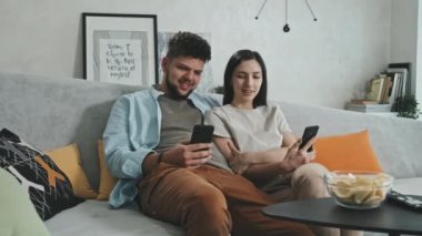 Genç Latin bir çiftin birlikte gülüp, akıllı telefonlarındaki komik medya içeriğini izlediği orta boy bir PAN fotoğrafı.